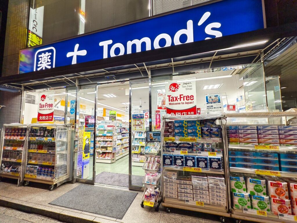Tomod's drugstore entrance in Japan at night showing tax-free shopping signage with cherry blossom logo, illuminated product displays, and "Welcome to Japan" message for international tourists