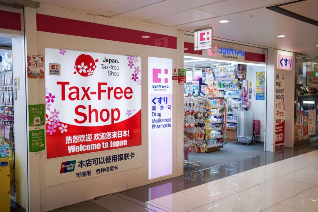 Tax-free shop storefront in Japan with red and white signage featuring cherry blossoms, welcoming international tourists with multilingual signs including "Welcome to Japan" and pharmacy products visible inside